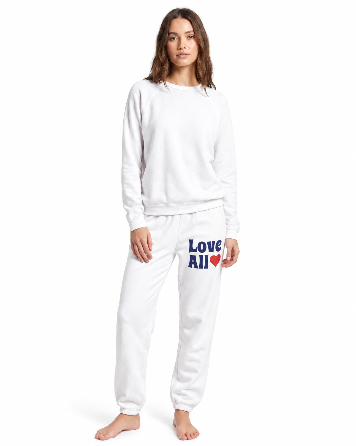 A woman modeling a white two-piece sweatshirt and jogger pants set. The sweatpants feature the text 'Love All' in blue with a red heart on the left thigh.
