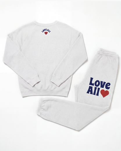 A white sweatshirt and matching sweatpants set laid flat on a white background. The sweatshirt has 'LOVE ALL' printed in blue with a small red heart on the upper back.