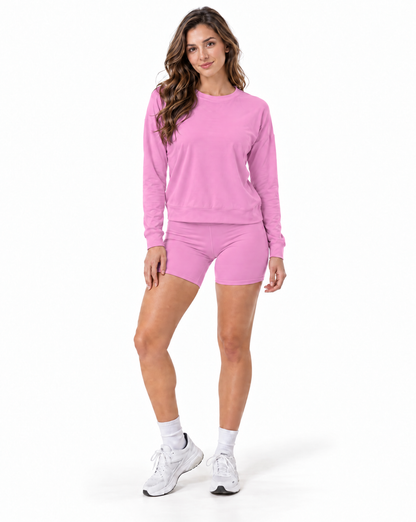 A woman modeling a matching pink activewear set consisting of a long-sleeve crewneck sweatshirt and matching athletic shorts, paired with white socks and white sneakers, against a plain white background.