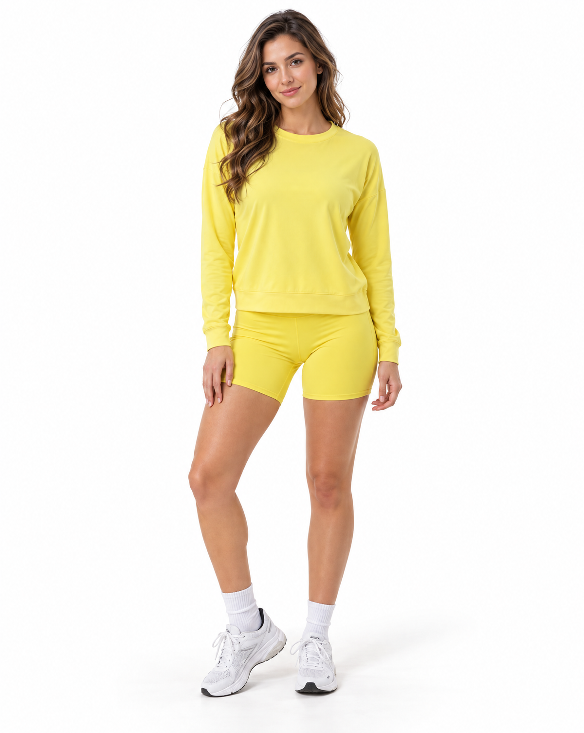 A woman modeling a matching bright yellow activewear set consisting of a long-sleeve crewneck sweatshirt and high-waisted biker shorts, paired with white socks and white sneakers, against a plain white background.