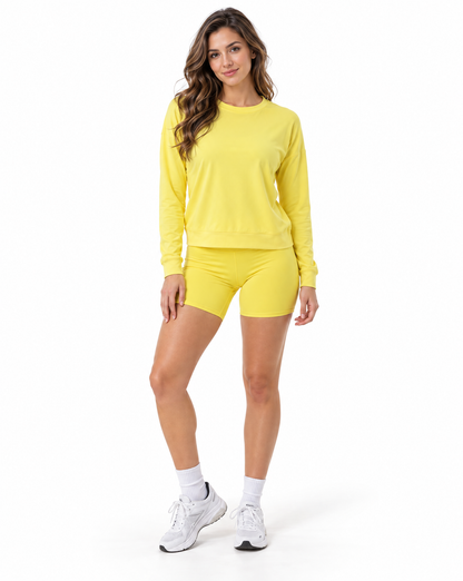A woman modeling a matching bright yellow activewear set consisting of a long-sleeve crewneck sweatshirt and high-waisted biker shorts, paired with white socks and white sneakers, against a plain white background.