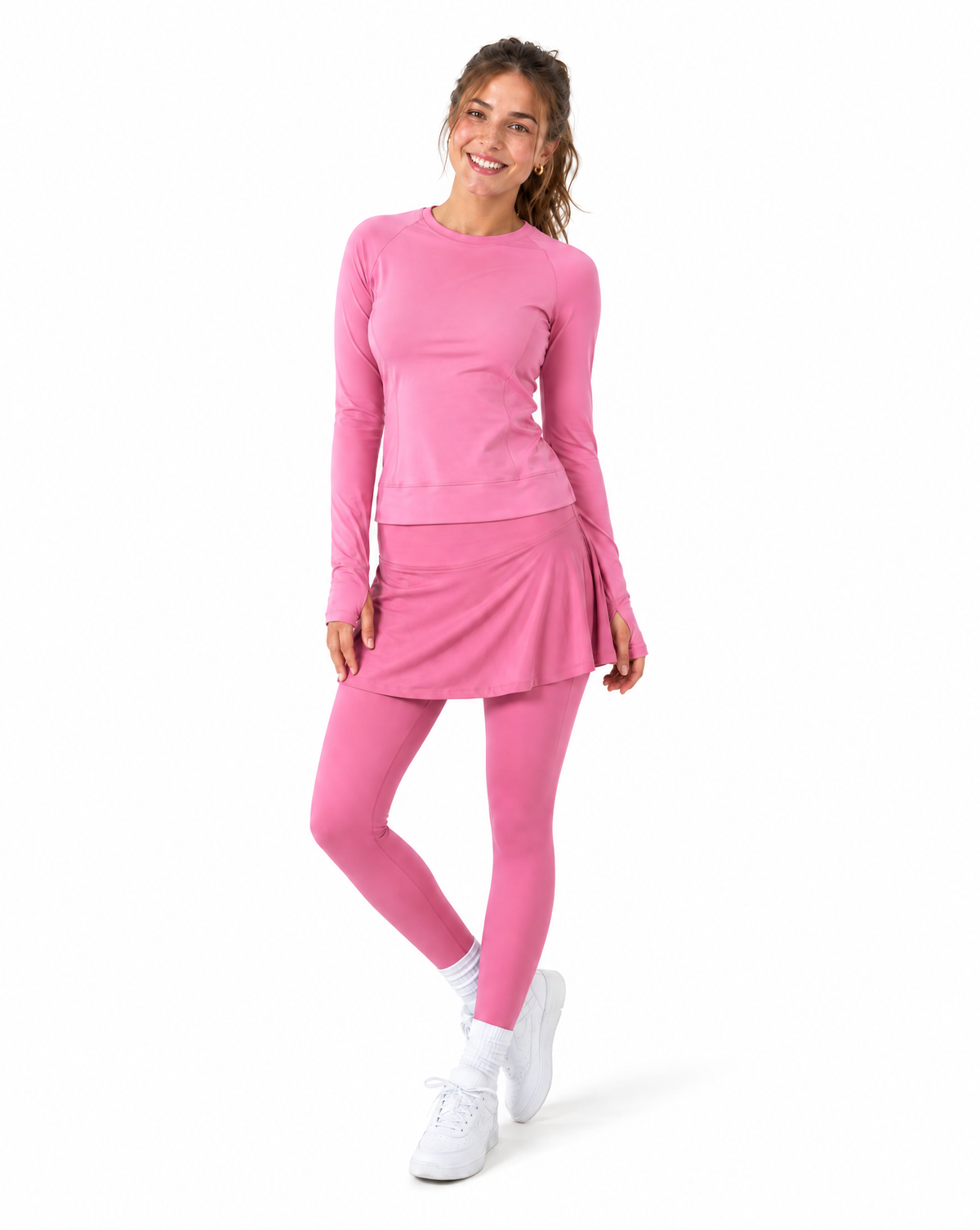 Woman wearing pink athletic long sleeve top and skirted leggings with sneakers set against a flat white background