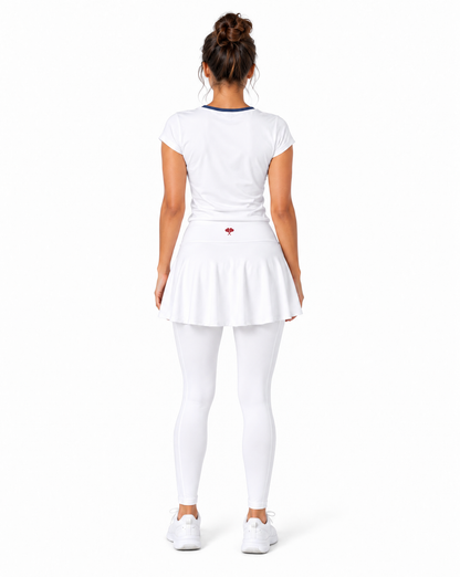 A woman viewed from the back wearing a white tennis outfit consisting of a short-sleeve top with a thin dark blue collar, a pleated tennis skirt with a small red heart logo on the back, white leggings, and white sneakers, set against a plain white backgro