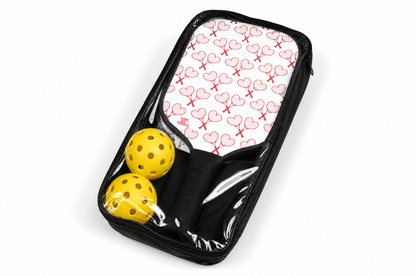 A black zippered carrying case containing a paddle racket set with a white and pink heart patterned paddle and two yellow perforated balls, shown on a white background.