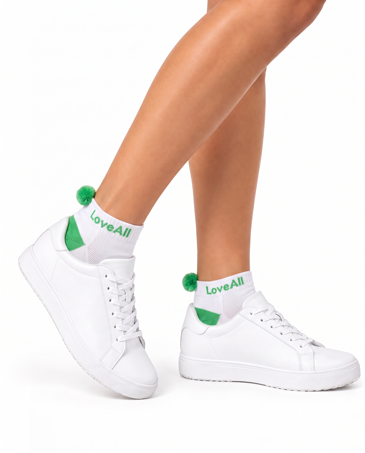 White low-top sneakers with green socks with green pom-poms, shown on a model's feet with white socks printed with 'LoveAll' on a clean white background.
