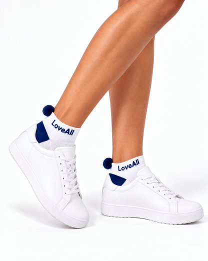 White low-top sneakers worn with white and navy pom pom socks printed with 'LoveAll', shown on a model's feet against a white background.