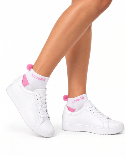 White low-top sneakers with pink heel tabs and white socks featuring the pink LoveAll logo, shown on a model's feet against a white background.