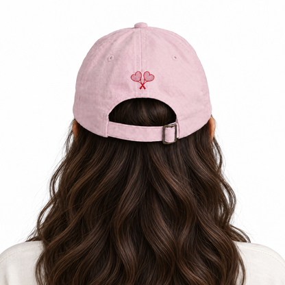 Back view of a woman with long wavy brown hair wearing a light pink baseball cap with a small red embroidered heart design on the front, set against a plain white background.