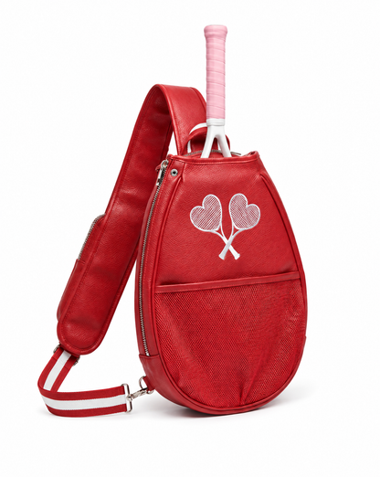 A red tennis bag with a textured leather-like finish, featuring a white embroidered crossed tennis rackets design on the front pocket. A pink tennis grip is tucked into the top opening, and a matching red and white shoulder strap is attached.