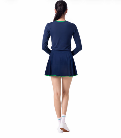 A woman shown from the back wearing a navy blue long-sleeve athletic top and skirt with a flared skirt and a thin green trim along the neckline and hem, paired with white socks and white shoes, against a plain white background.