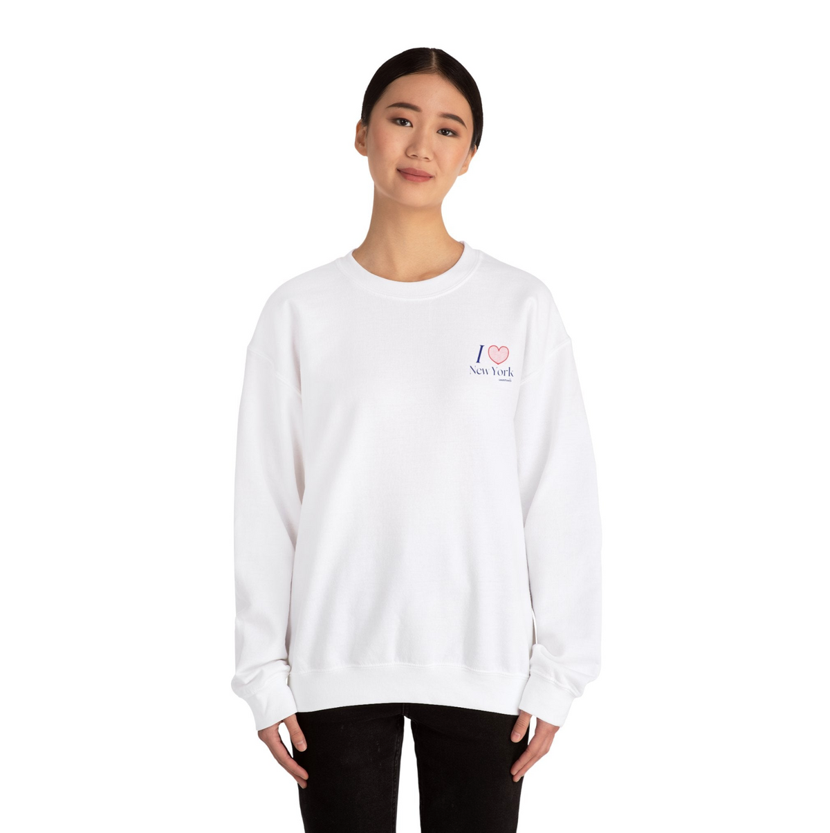 Woman wearing a white sweatshirt with a "I heart New York" graphic on the left hand side of the chest with black pants on white studio background