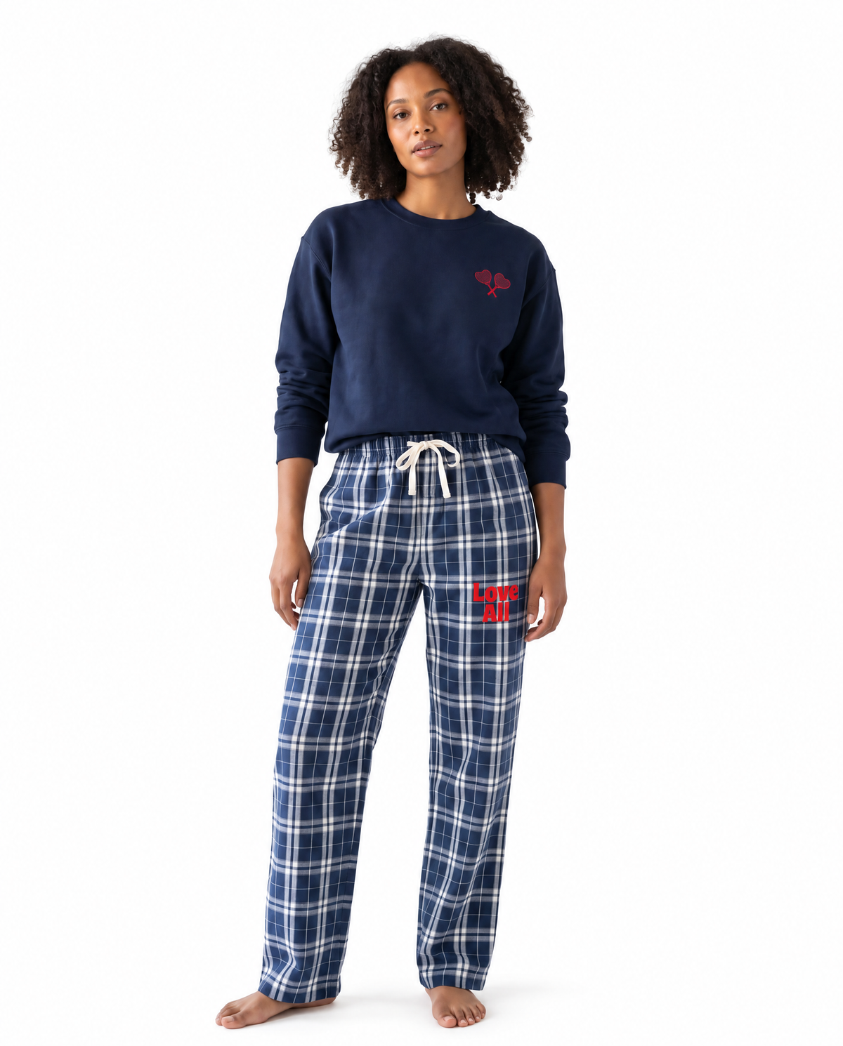 A woman modeling a navy blue crewneck sweatshirt and matching navy plaid pajama pants with a red embroidered logo on the chest and thigh, set against a plain white background.