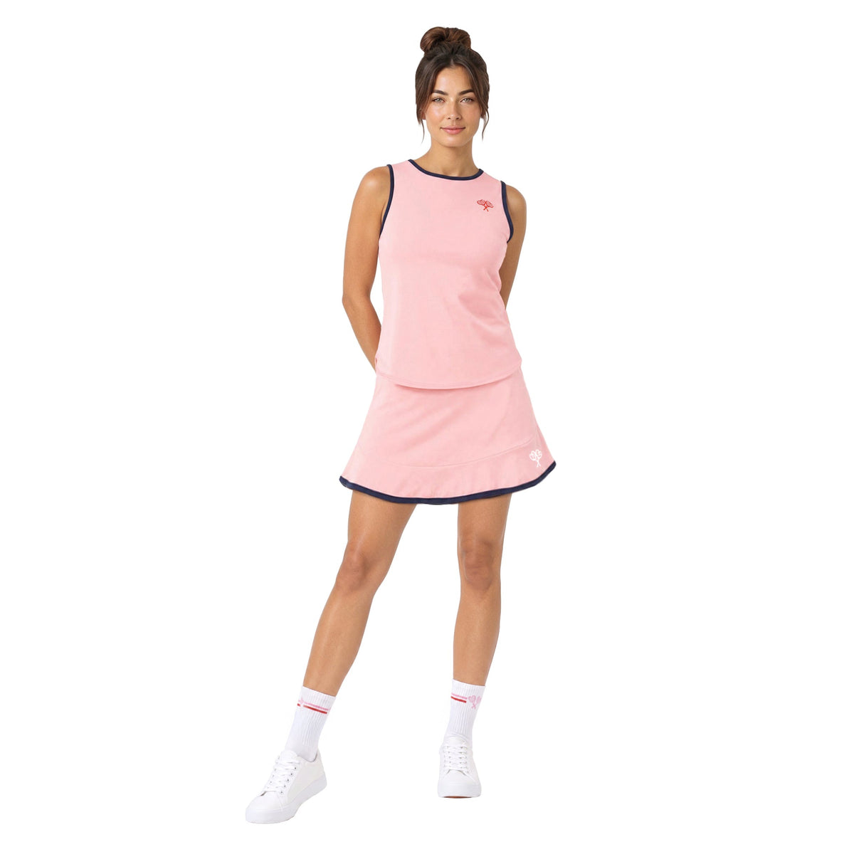 A woman modeling a light pink sleeveless tennis dress with dark navy trim, paired with white socks and white sneakers, against a solid white background.