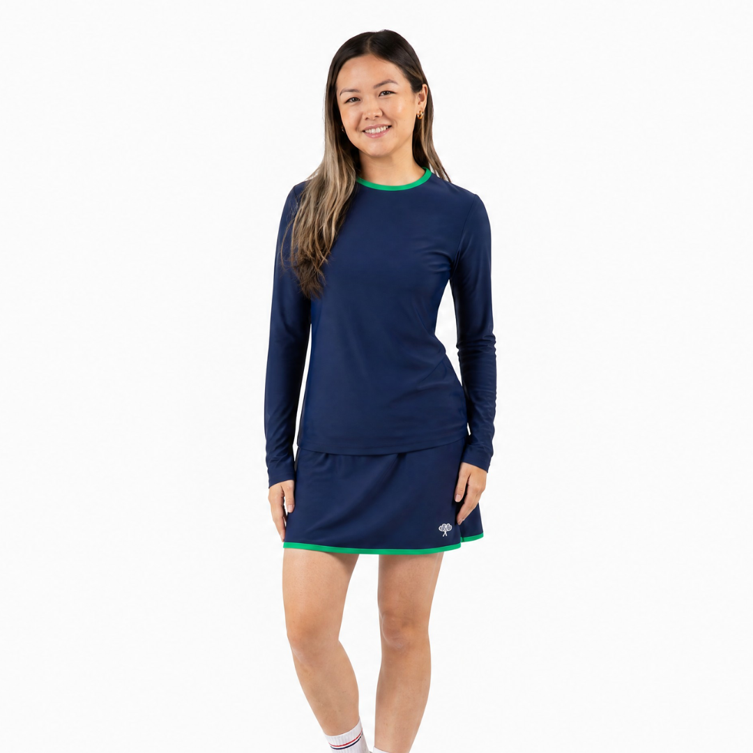A young woman modeling a navy blue long-sleeve athletic skort and top with green trim at the crew neck and hem, paired with white socks and white sneakers, against a solid white background.