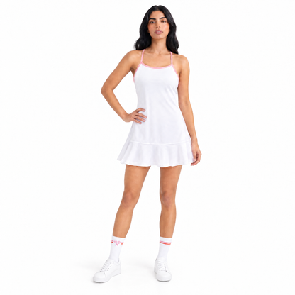 A woman modeling a white sleeveless tennis dress with thin pink straps and a ruffled hem, paired with white socks and white sneakers, against a plain white background.