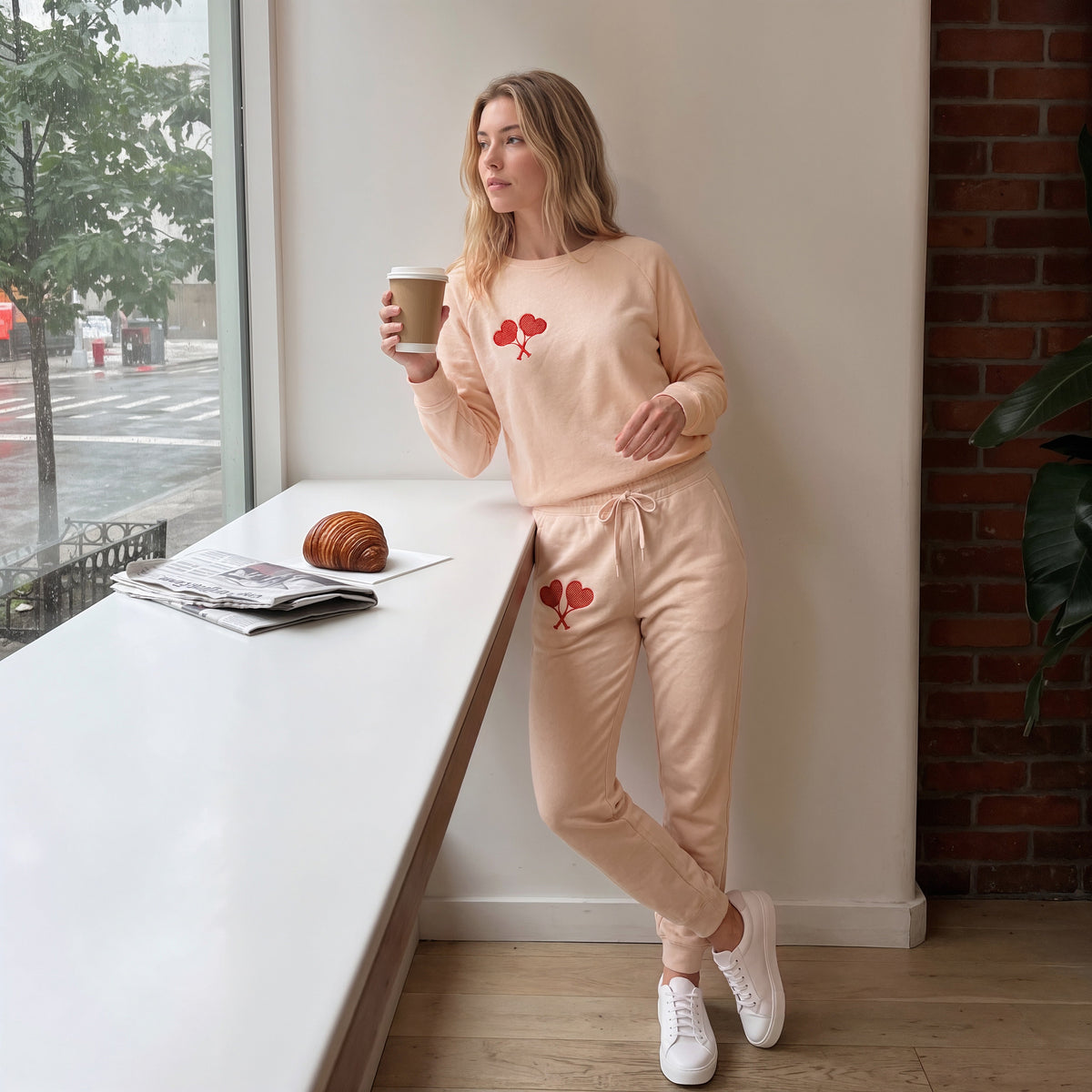 A woman stands by a window in a bright, modern room wearing a light peach two-piece lounge set with red heart graphics. She holds a coffee cup, with a newspaper and croissant on the white windowsill next to her.