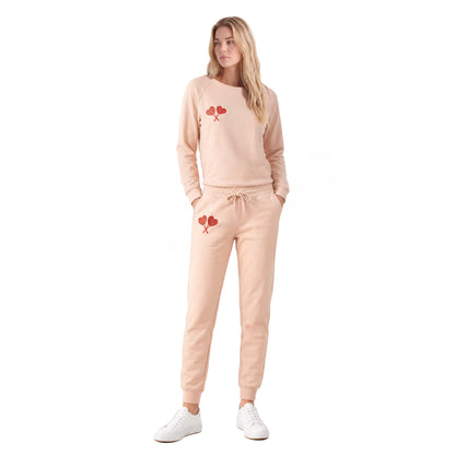 A woman modeling a light peach-pink sweatshirt and jogger sweatpants set with red heart graphics, paired with white sneakers, on a solid white background.