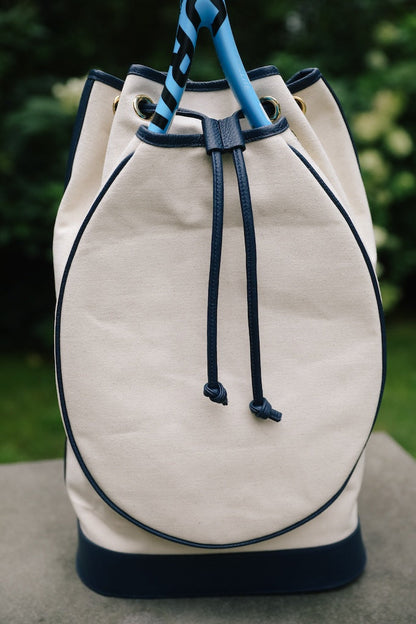An off-white canvas bucket bag with navy blue trim, drawstring closure, and a blue-and-white striped top handle, photographed outdoors on a stone surface with blurred green foliage in the background.