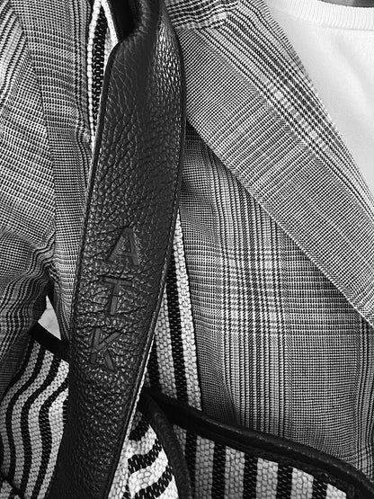 Close-up of a black textured leather shoulder strap with the brand name 'ATK' embossed vertically, worn over a black-and-white plaid blazer and striped garment.