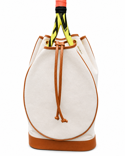 A canvas bucket bag in off-white with tan leather trim, featuring a drawstring closure and a tennis racket tucked inside, set against a plain white background.