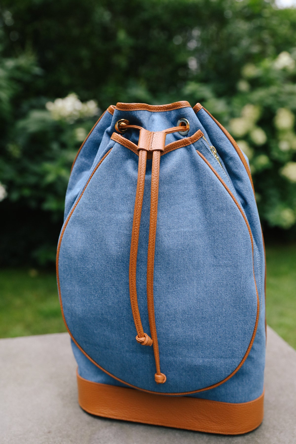 A blue canvas drawstring backpack with tan leather trim and base, displayed on a stone surface with a blurred garden background.