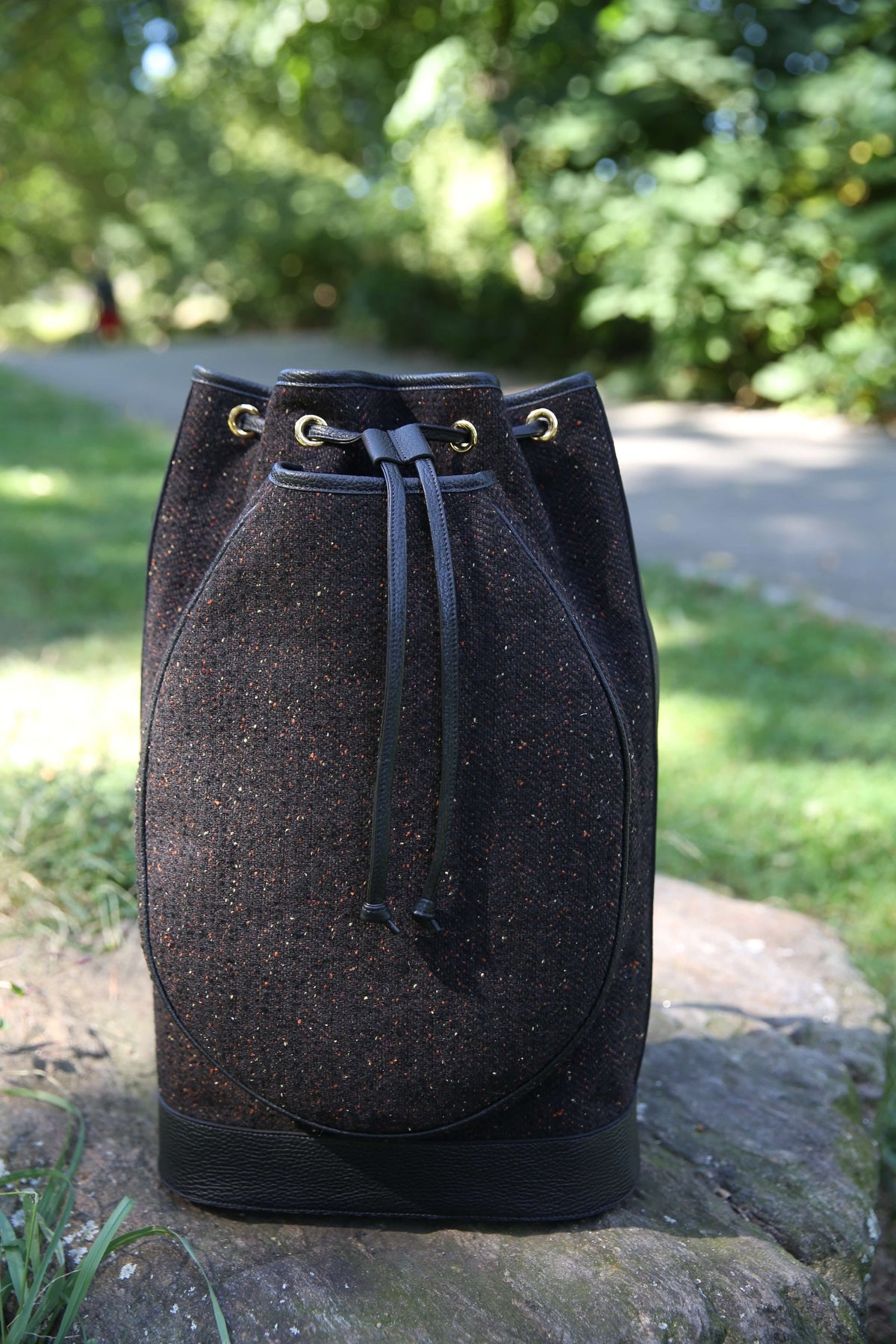 A black textured bucket bag with black drawstrings and gold-tone eyelets, resting on a stone in an outdoor park setting with green grass and trees in the background.