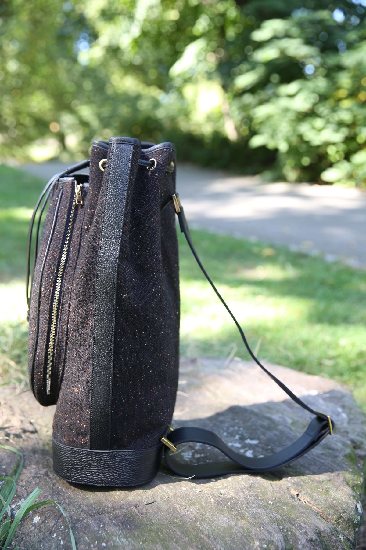 A black textured bucket bag with black leather trim and a long shoulder strap, resting on a stone in an outdoor garden setting.