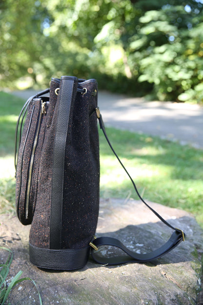 A black textured bucket bag with black leather trim and a long shoulder strap, resting on a stone in an outdoor garden setting.