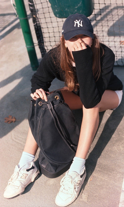 A person sitting outdoors on a concrete surface near a fence, wearing a black New York Yankees baseball cap, a black long-sleeve top, white shorts, white socks, and white sneakers, while holding a large black bucket bag.