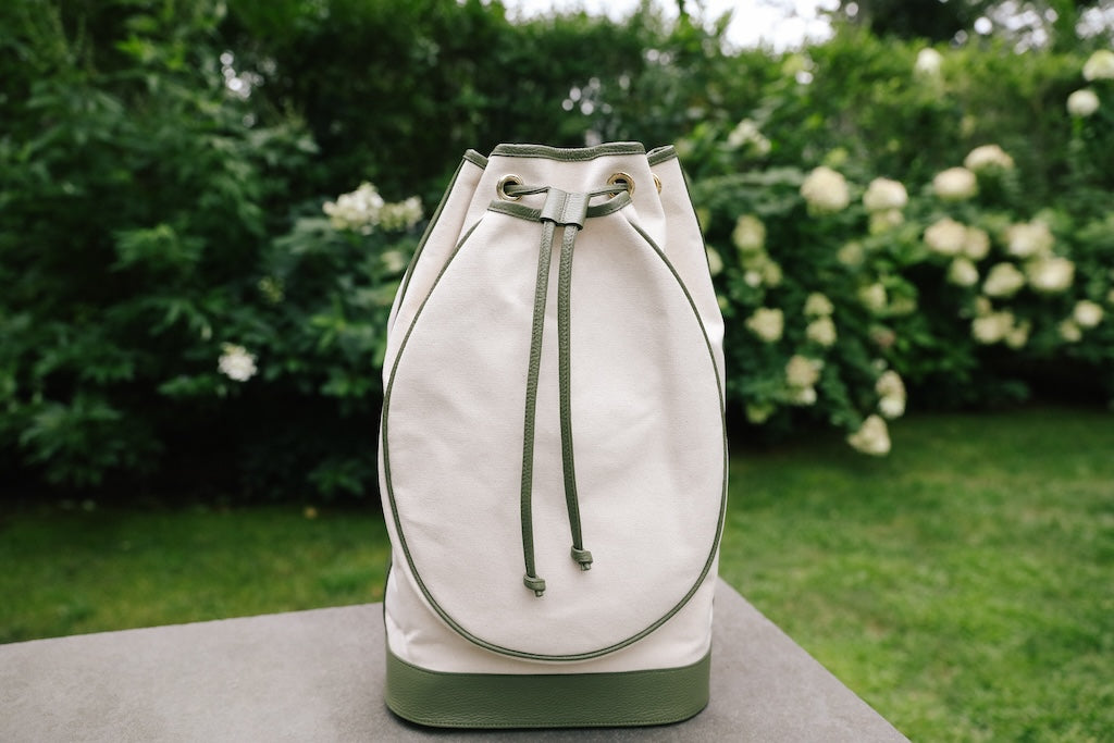 A cream canvas drawstring bucket bag with sage green trim and base, displayed on a stone surface with a blurred garden background.