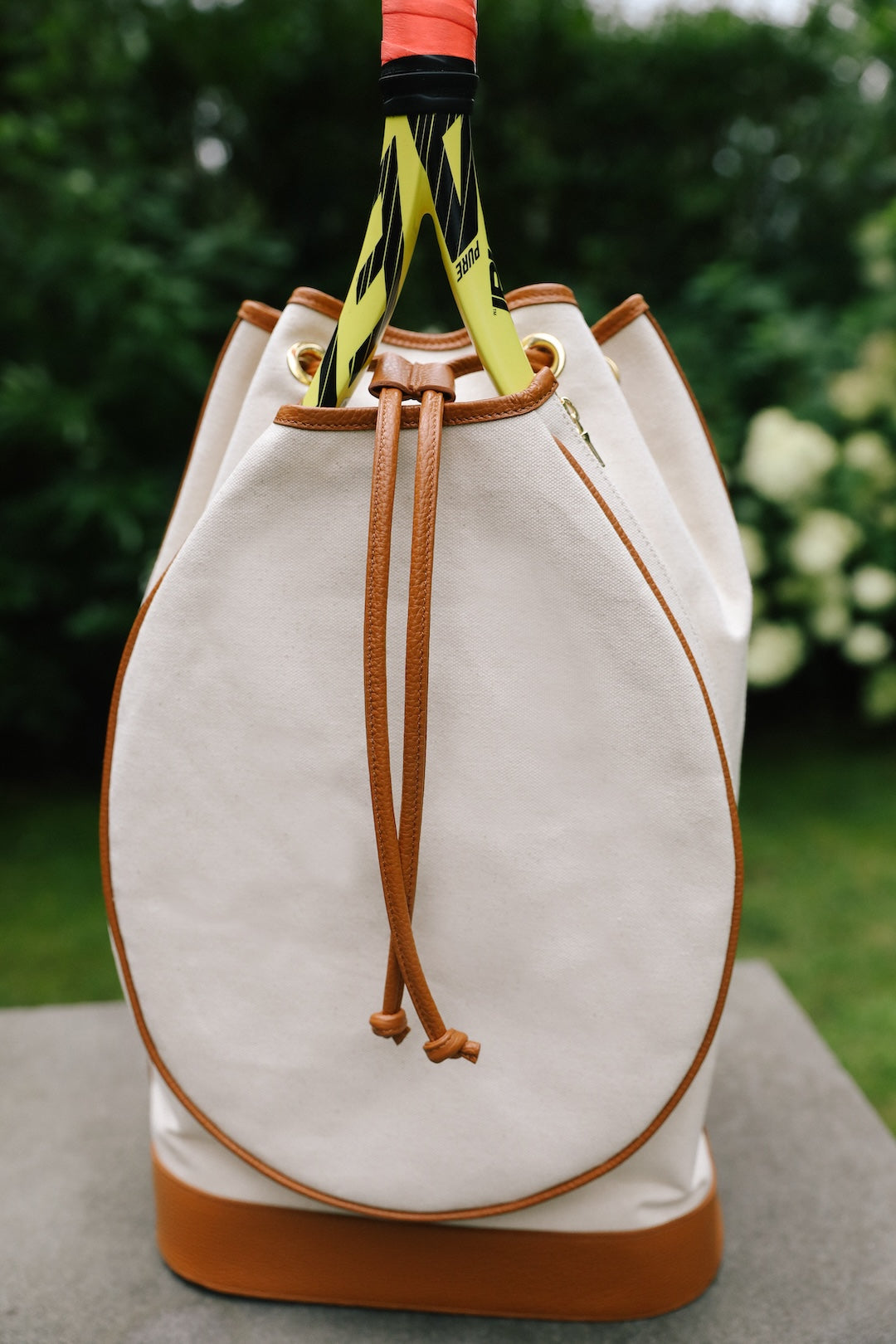 A white canvas tennis bag with tan leather trim and drawstring closure, featuring a tennis racket handle wrapped in a yellow and black striped grip tape, set against a blurred green garden background.
