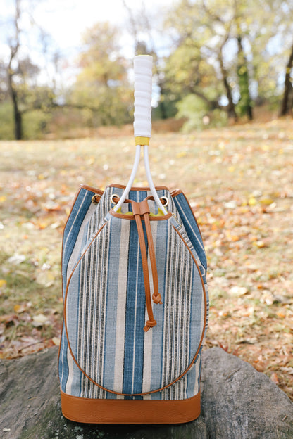 A striped bucket bag with blue, beige, and white vertical stripes, tan leather trim, and a white wrapped handle, sitting on a stone in an outdoor park setting with fallen leaves.