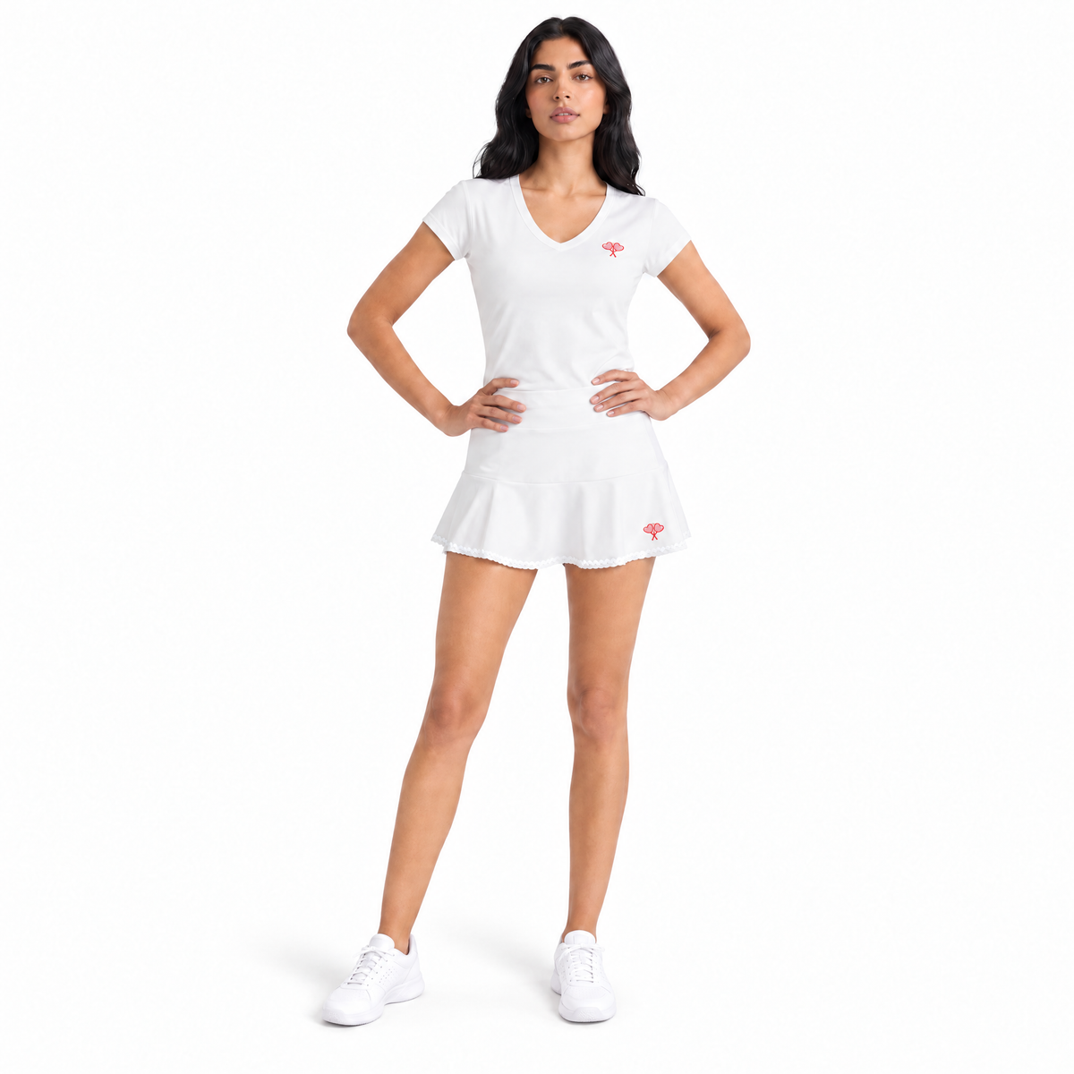 A woman modeling a white short-sleeve V-neck tennis dress with a ruffled hem, featuring small coral logos on the chest and skirt, paired with white sneakers against a solid white background.
